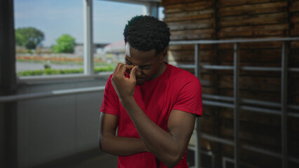 Man with hand on chin in building interior near window and railing wearing red shirt with furrowed...