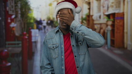 Man in red shirt and santa hat covers eyes with hand amid blurred pedestrians on busy city street; shyness.