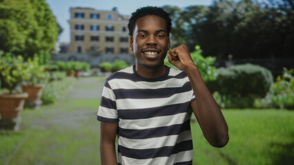 Naklejka premium Young african american man in striped shirt smiles and raises fist on paved park walkway beside planters; empowerment.