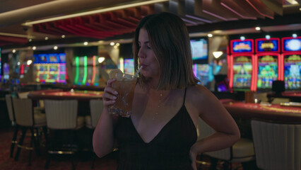 Young brunette woman holding cocktail glass and smiling while toasting near slot machines in casino...