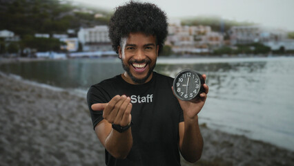 Smiling man with afro hair in staff uniform holding a clock, extends his hand towards the camera with a seaside backdrop of sand, water, and buildings in view.