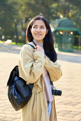 Obraz premium Traveler with camera and backpack in a sunlit park, casual coat and scarf, ready for street photography and urban exploration under warm light, curious expression.
