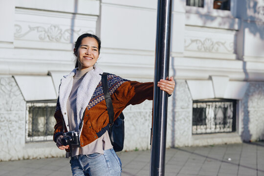 Smiling woman holds a pole and a camera, wearing a patchwork jacket and jeans, photographed outdoors on a city street during casual photography exploration.