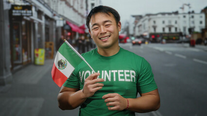Young man in green volunteer shirt holds mexican flag on urban street showcasing cultural diversity outdoors in city setting with smiling expression.
