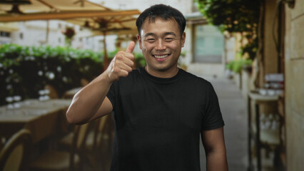 Fototapeta premium Man smiling and giving thumbs up gesture on street cafe terrace wearing black t shirt under bright sunlight; approval.