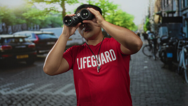 Man wearing red lifeguard shirt holds binoculars and points finger upward on busy street; alertness.
