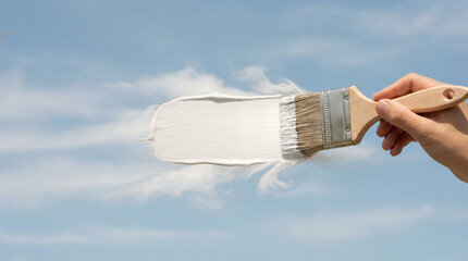 Fototapeta premium A hand with a paintbrush applies a white stroke on a blue sky with clouds. Creative concept for the color of the year 2026 trend. Imagination and new beginnings