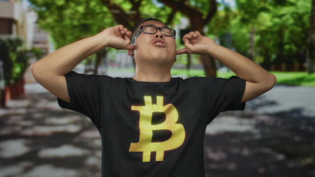 Young man plugging fingers into ears, squinting with closed eyes while wearing a black bitcoin t shirt on a tree lined street; crypto frustration. - Powered by Adobe