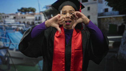 Woman wearing graduation gown and cap shows thumbs down gesture at port dock beside moored boat...