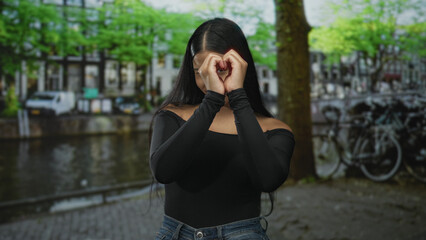 Fototapeta premium Woman makes heart sign with hands by canal under leafy trees and winks toward camera; affection romance connection.