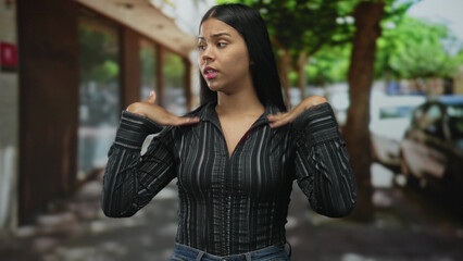 Woman in striped shirt fans bare hands on a sunlit city street lined with trees and parked cars in afternoon heat; heat discomfort.