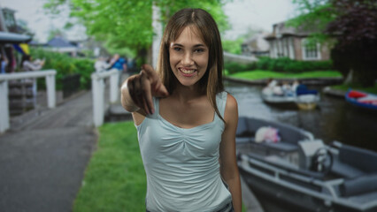 Young woman points finger at camera on a street in amsterdam wearing a light blue tank top and jeans, canal boats behind her; playful invitation.