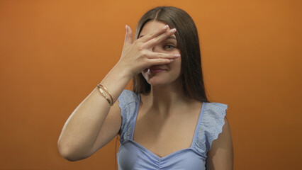 Teenage girl covers eyes with hand in studio against orange wall; vulnerability introspection...