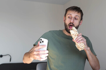 A man takes a selfie while eating a large wrap, capturing a humorous moment of multitasking with fast food at home. Casual lifestyle scene reflecting modern habits and social media behavior.