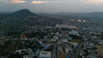Aerial view of Pushkar Lake during the fair, capturing cloud reflections on the water, glowing roller-coaster lights, lakeside houses and ghats, with a warm sunset over the Aravali hills in the back