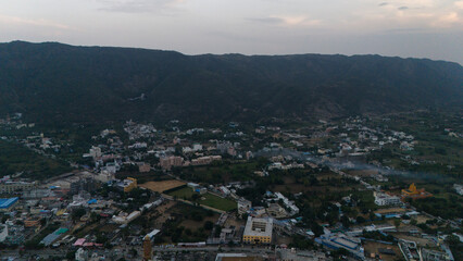 Aerial pan shot of Pushkar town at sunset, showing houses, Pushkar Lake, glowing evening light, cloudy sky, and the Aravali hills creating a scenic backdrop across the serene landscape from above.
