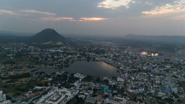 Aerial view of Pushkar Lake during the fair, capturing cloud reflections on the water, glowing roller-coaster lights, lakeside houses and ghats, with a warm sunset over the Aravali hills in the back