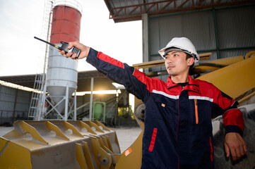 An industrial engineer directing operations at a concrete batching plant, communicating via walkie-talkie near heavy machinery. Professional supervision ensuring safe production workflow.