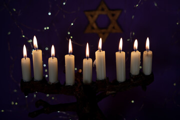 A menorah with nine glowing candles is placed in front of a Star of David on a purple background, representing Hanukkah.