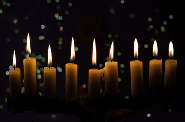 A menorah with nine glowing candles is placed in front of a Star of David on a purple background, representing Hanukkah.