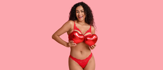 Beautiful young African-American woman in stylish red lingerie with air balloons in shape of heart on pink background. Valentine's Day celebration