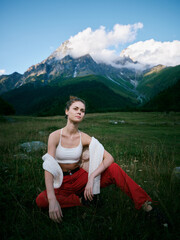 Fototapeta premium Woman sits in a grassy field with a mountain backdrop, wearing a white crop top and red pants, relaxed pose in natural light, outdoor scenery and serene alpine landscape