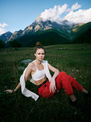 Fototapeta premium Woman in white top and red pants sits in a meadow with towering mountains and clouds, outdoor fashion and nature inspiration for travel, leisure, and adventure photography