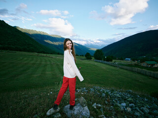 Fototapeta premium Woman in countryside field wearing whitesweater and redpants smiling near mountains under blue sky outdoors moment showcasing nature meadow rural scenery