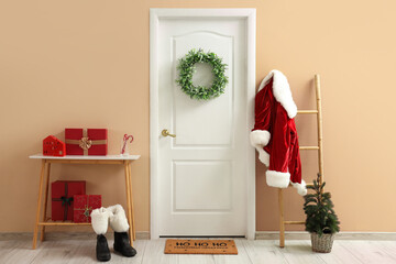 Fototapeta premium Christmas home entrance with white door, green wreath, Santa costume on ladder, gift boxes, boots and festive decor in cozy minimalist interior