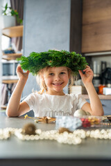 Naklejka premium Little girl wearing a Christmas wreath on her head and laughing during holiday decorating. Festive joy, family traditions, and playful moments during Christmas preparation.