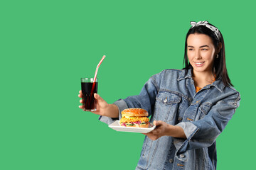 Beautiful young woman holding plate with tasty burger and glass of cola on green background