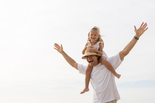 Happy father carrying daughter on shoulders at the beach, both smiling and enjoying summer vacation, representing fatherhood, family bonding, parenting joy and playful lifestyle in tropical setting.
