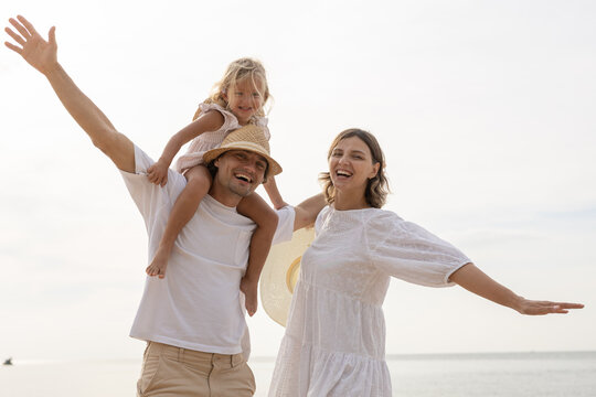 Cheerful family enjoying time together at the beach, with daughter on father shoulders and mother smiling beside, symbolizing love, bonding, parenthood and joyful summer vacation lifestyle.