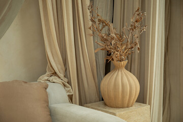 A stylish wooden vase holds dried flowers next to soft curtains