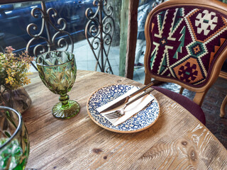 Cozy restaurant table setting with patterned plate, cutlery on a napkin, green glassware, and an ethnic-style chair, creating a warm and inviting dining atmosphere with rustic wooden textures.