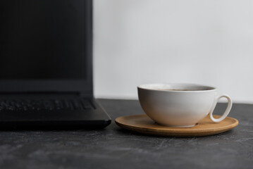 A white coffee cup on a wooden saucer next to a black laptop on a dark surface. The scene suggests a cozy workspace or coffee break.