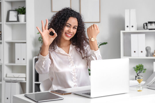 Young African-American businesswoman with wristwatch showing OK at table in office. Time management concept - Powered by Adobe