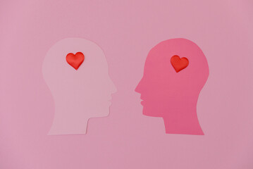 Two paper cutouts of human heads in profile, one white and one pink, each with a red heart shape in the forehead area, set against a pink background.