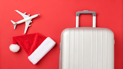 Travel items arranged on a red surface for holiday trips with a suitcase, airplane model, and Christmas hat