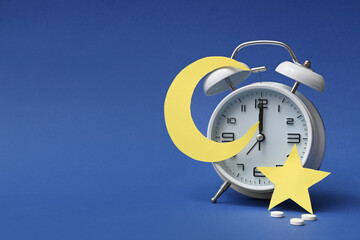 Alarm clock with sleeping pills, paper star and moon on blue background. Melatonin concept