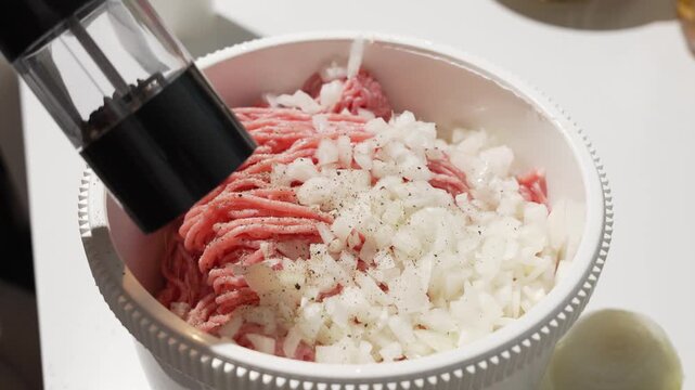 Freshly ground black pepper falls onto minced meat and chopped onions in a bowl. Seasoning step for burgers, meatballs, or sausages.