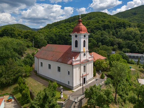 Aerial view of the Church of St. Luke with its red roof and white walls, nestled against a backdrop of green hills, Sklene Teplice, Banska Bystrica Region, Slovakia.