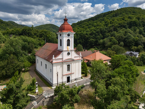 Aerial view of St. Luke's Church standing proudly with its red roof and white walls amidst a sea of green trees under a cloudy sky, Sklene Teplice, Banska Bystrica Region, Slovakia.