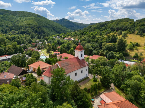 Aerial view of the Church of St. Luke nestled among lush green trees and houses with red roofs, under a sky of scattered clouds., Sklene Teplice, Banska Bystrica Region, Slovakia.