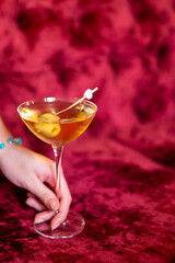 A hand holds a classic amber cocktail in a martini glass, garnished with olives, against a luxurious red velvet background