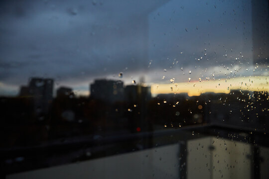 Dark and moody cityscape view through a window covered in raindrops at dusk, highlighting the blurred skyline silhouettes against a dramatic, colorful sky.