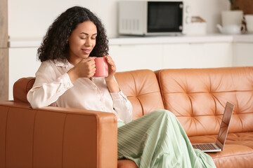 Beautiful young African-American businesswoman with cup of hot coffee and modern laptop sitting on sofa at home