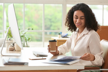 Beautiful young African-American businesswoman working with documents and paper cup of hot coffee at table in office