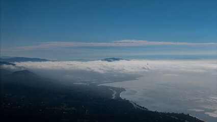 Aerial view of the coastline where the dark, forested hills meet the bright, reflective ocean, partially obscured by a layer of dreamy, low-lying clouds, Montecito, California, United States.