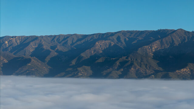 Aerial view of mountains pierce through a sea of ethereal fog under a clear blue sky, creating a serene yet dramatic landscape, Montecito, California, United States.